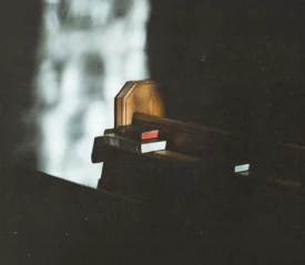 A wooden church pew is lit by a sliver of light, with hymnals and a Bible tucked into the rack behind it