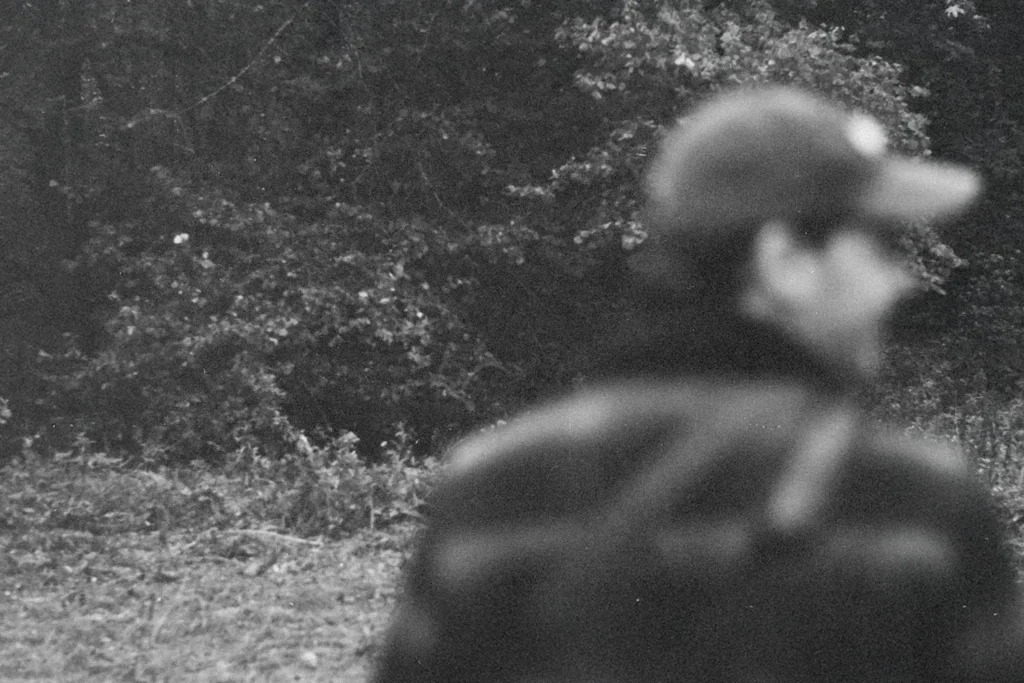 Man wearing a hat, standing in a wooded clearing with dense trees in the background
