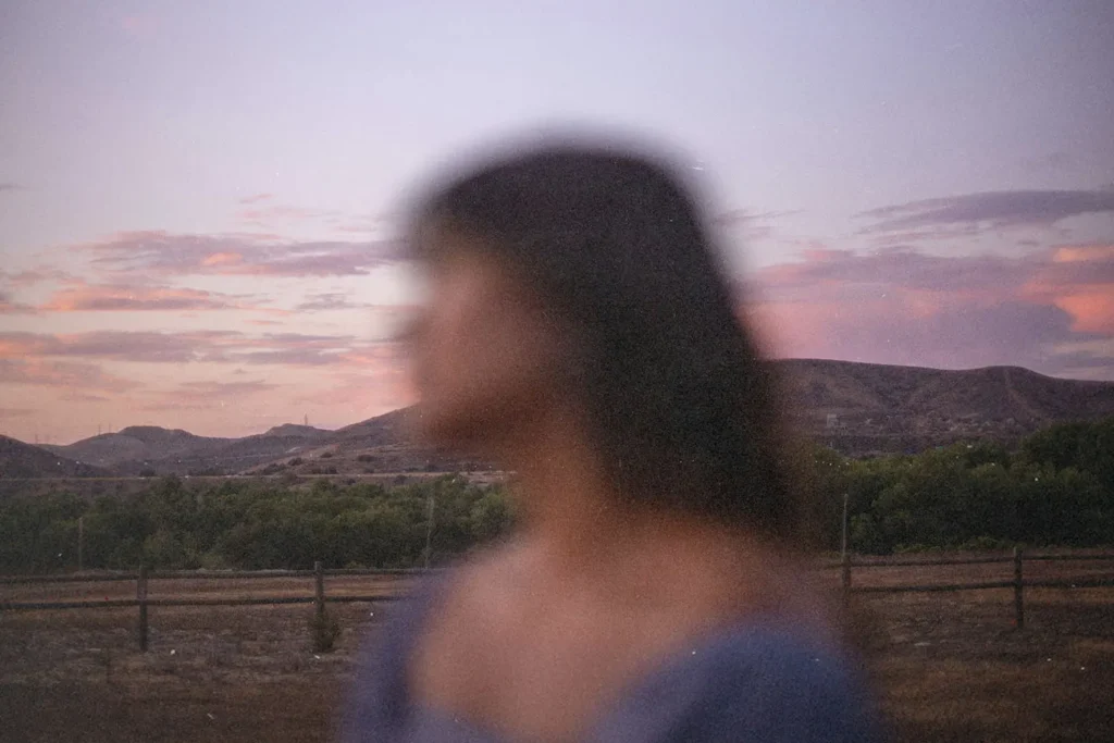 Out-of-focus person positioned before a rural landscape featuring hills, trees, and a pink and purple sunset sky