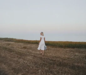 A woman in a white dress walks through a dry, open field near the shoreline under a pale evening sky