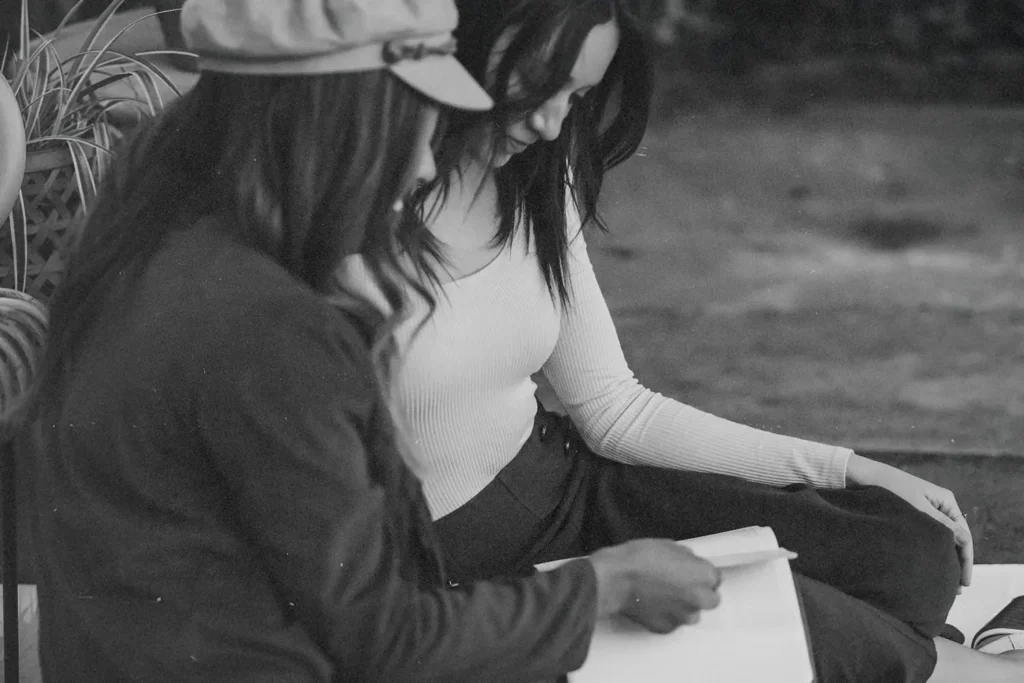 Two people sitting together outdoors, reading and discussing an open book in a relaxed setting