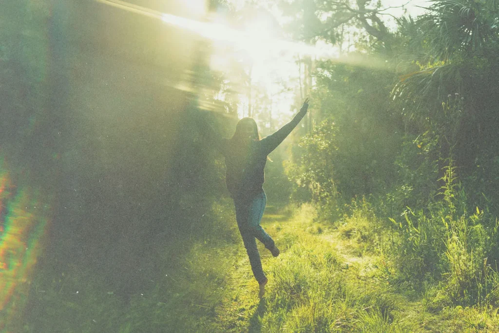 A person stands on a sunlit forest path with arms raised, surrounded by tall trees and bright rays of light streaming through the foliage