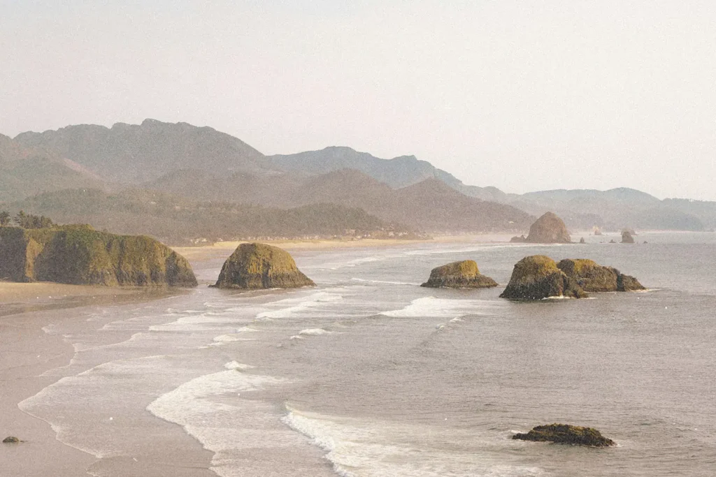 Ocean waves roll toward a sandy shoreline with large rocky sea stacks and distant mountains under a hazy sky