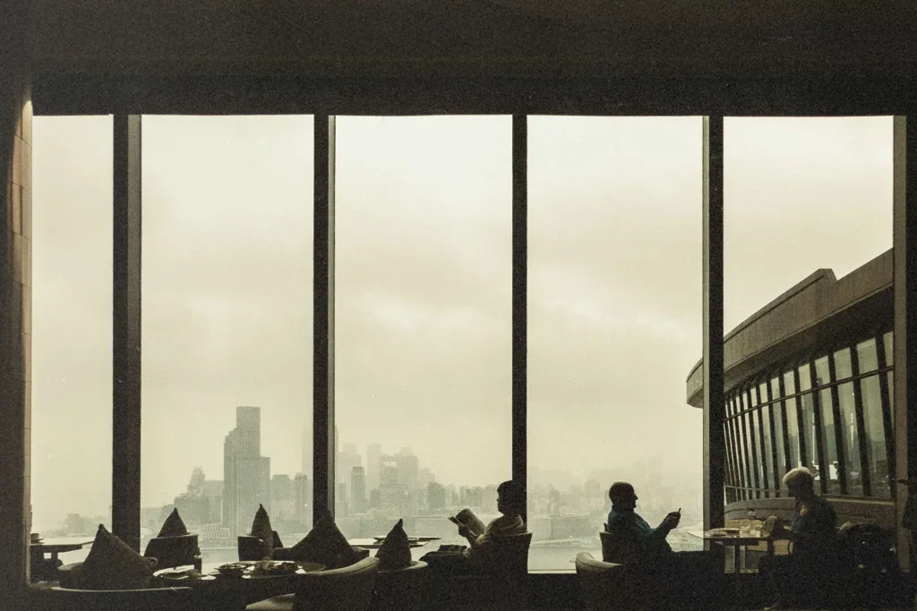 People sit at tables inside a restaurant with tall windows overlooking a cloudy city skyline