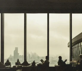 People sit at tables inside a restaurant with tall windows overlooking a cloudy city skyline