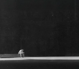 Person sitting alone on a long bench in a vast, dimly lit space, leaning forward with their head down