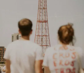 Two people stand outdoors looking toward a tall red metal tower, with buildings and a crowd in the background