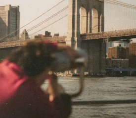 A person looks through a public viewing telescope toward a large suspension bridge crossing over the water
