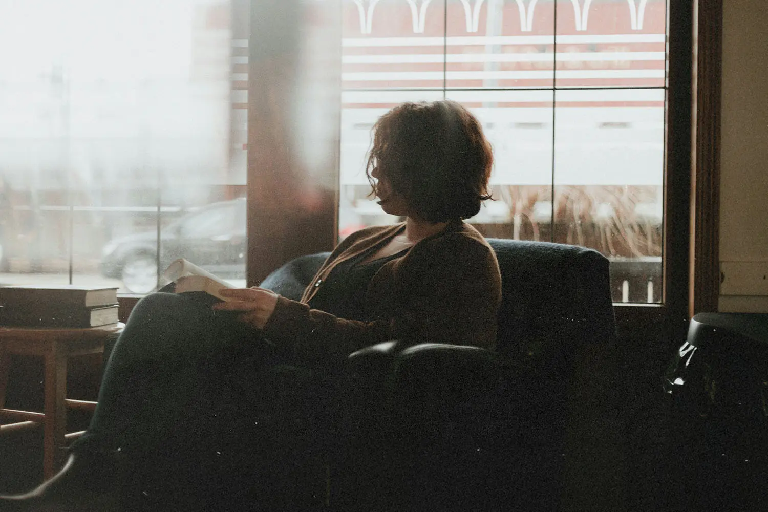 A person sits in a cozy chair near a large window, reading a book as soft daylight filters into the room