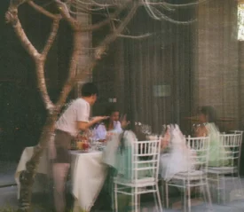 Server attending to a group of people dining at an outdoor restaurant, seen through a glass window with soft motion blur