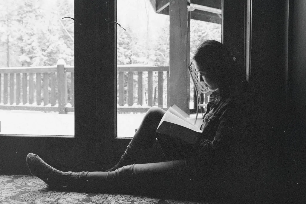 A person sits on the floor beside large glass doors, reading a book while snow-covered trees and a porch are visible outside