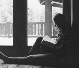 A person sits on the floor beside large glass doors, reading a book while snow-covered trees and a porch are visible outside