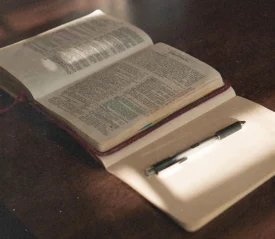 An open Bible rests on a table beside a blank notebook and pen, lit by soft sunlight from a nearby window
