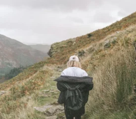 A person walks alone along a narrow mountain trail, wearing a light jacket and backpack as misty hills rise on both sides