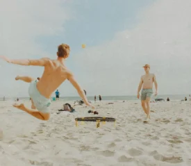 Two people on a beach playing spike ball, as one jumps into the air to hit the ball over the sand on a sunny shoreline