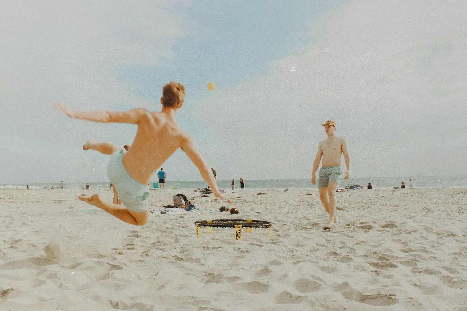 Two people on a beach playing spike ball, as one jumps into the air to hit the ball over the sand on a sunny shoreline