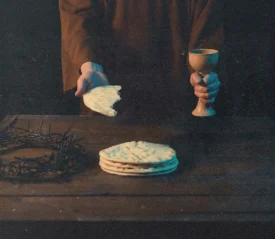 Hands holding unleavened bread and a communion cup above a wooden table with matzo and a crown of thorns