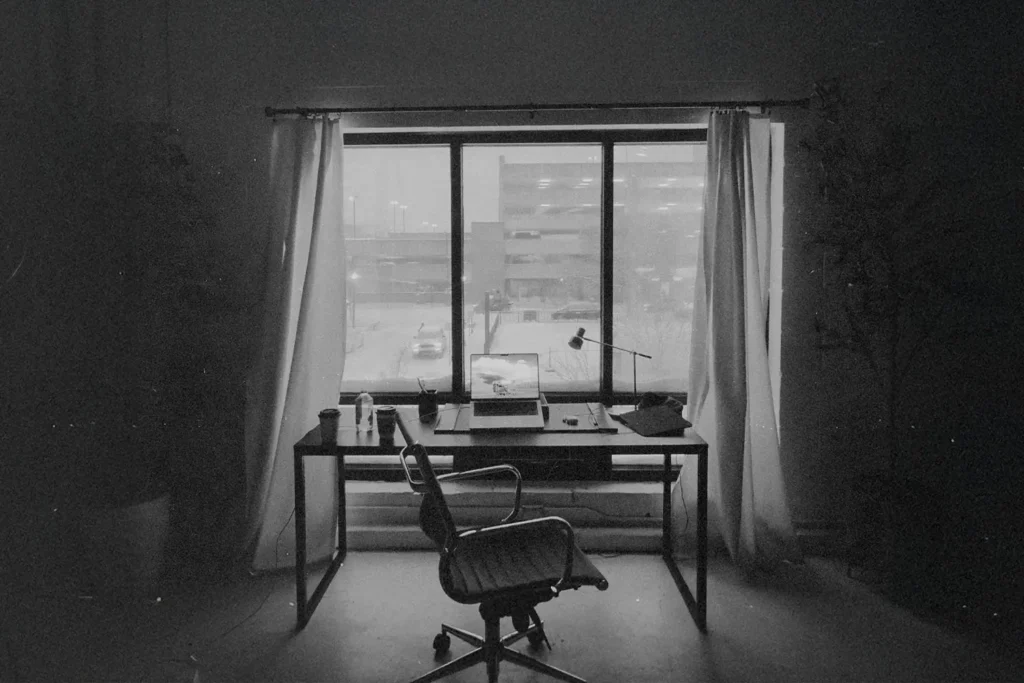 Simple office space with a laptop and lamp on a desk framed by open curtains and an urban view
