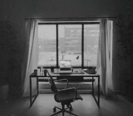 Simple office space with a laptop and lamp on a desk framed by open curtains and an urban view