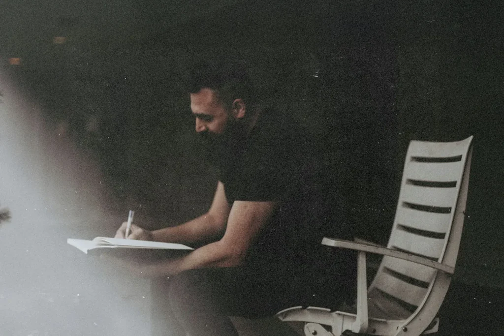 Person sitting in a chair and writing in a notebook, surrounded by soft, low lighting and a dark background