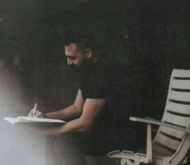 Person sitting in a chair and writing in a notebook, surrounded by soft, low lighting and a dark background