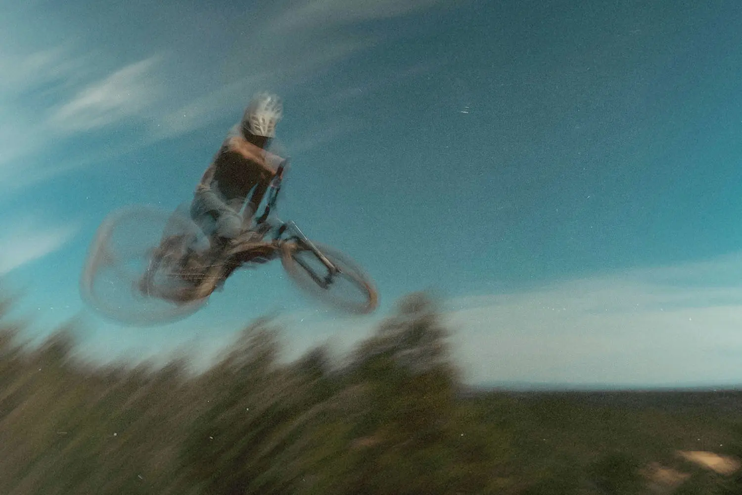 A cyclist performs a high jump on a mountain bike, captured in a motion‑blurred scene against a blue sky