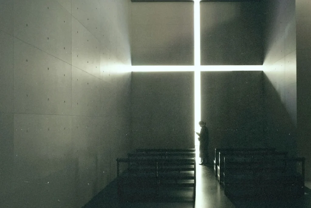 A person stands inside a dimly lit chapel, silhouetted against a large glowing cross-shaped opening on the far wall