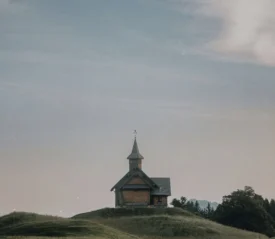 A small wooden chapel with a steeple sits alone on rolling green hills under a soft, pastel sky