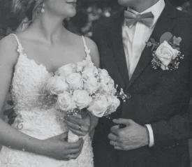 A bride and groom stand close together, the bride holding a bouquet of roses while the groom adjusts his suit jacket