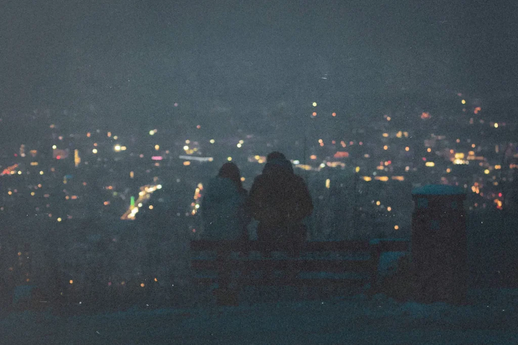 Two people sit on a bench at night overlooking a city filled with glowing lights in the distance