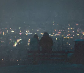 Two people sit on a bench at night overlooking a city filled with glowing lights in the distance