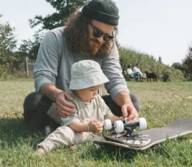 Father helping a baby examine a skateboard while sitting on the grass