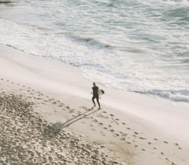 Surfer walking along the shoreline with a board under one arm, leaving footprints in the sand as waves roll in