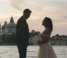 Married couple stand facing each other near the water at dusk, with historic domed buildings in the background and soft pastel colors in the sky