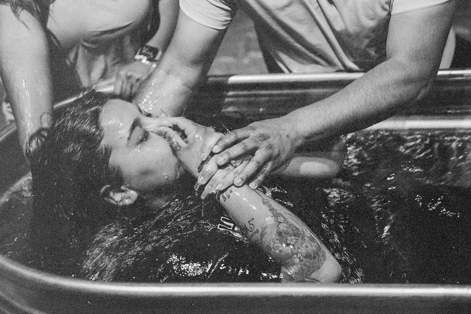 Two people assisting a woman being lowered into a water-filled baptism tub during a baptism ceremony