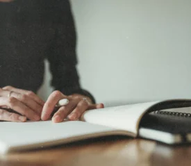 A person writes in an open notebook on a wooden table, with hands resting gently on the page