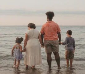 A family of four stands together at the edge of the ocean, holding hands as gentle waves wash over their feet during a calm sunset