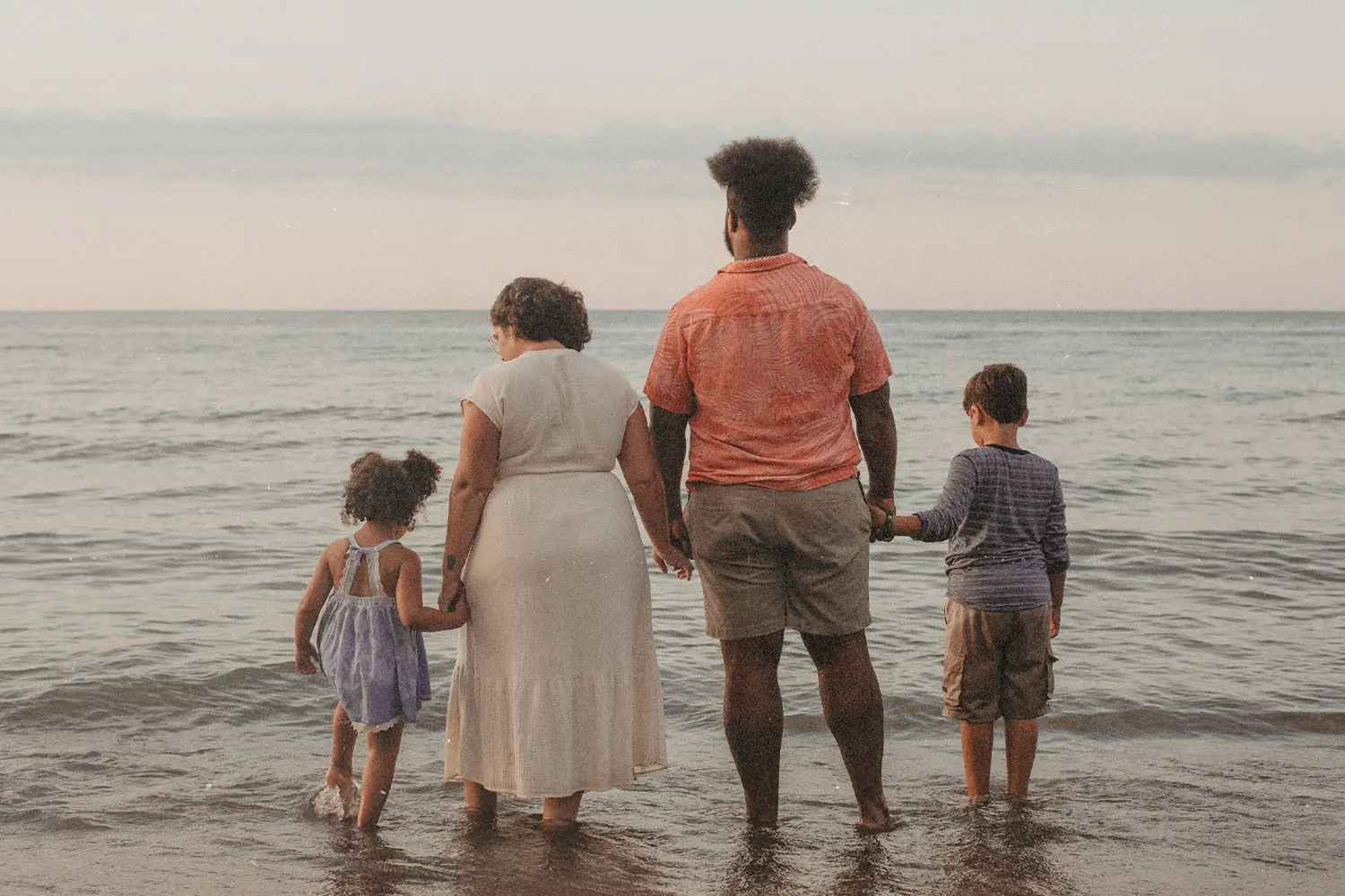 A family of four stands together at the edge of the ocean, holding hands as gentle waves wash over their feet during a calm sunset