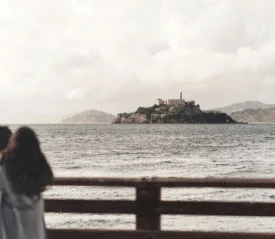 Mother holding her child, standing on a wooden pier looking across the water toward an island with historic stone buildings