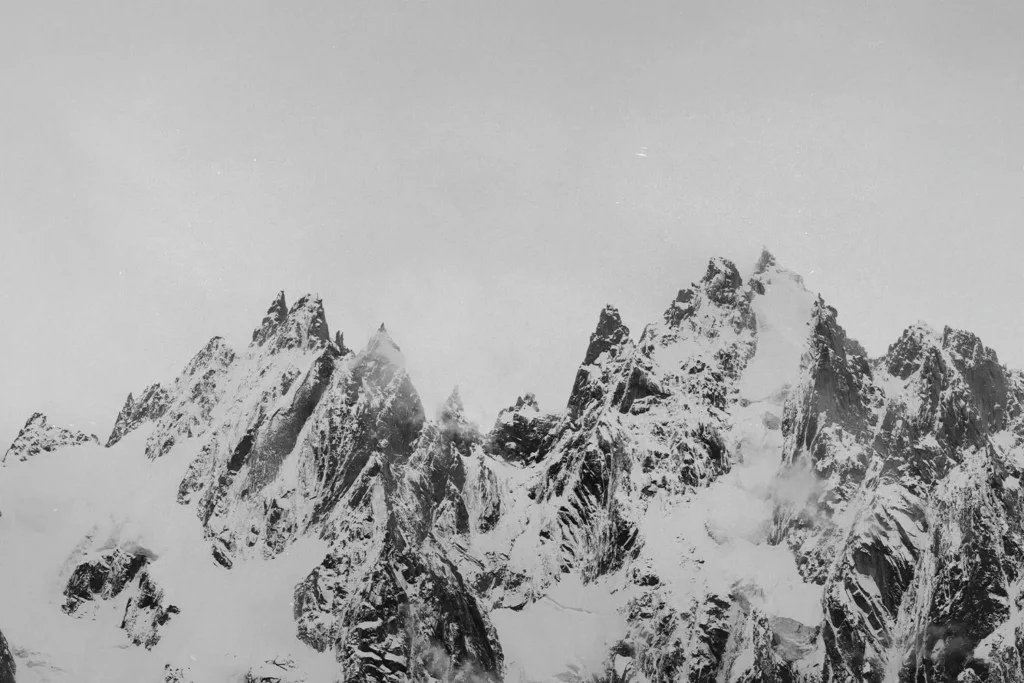 Sharp, jagged mountain peaks covered in snow rise dramatically into a pale, overcast sky, with patches of mist drifting around the ridges