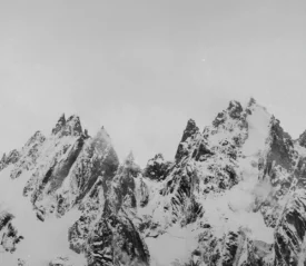 Sharp, jagged mountain peaks covered in snow rise dramatically into a pale, overcast sky, with patches of mist drifting around the ridges
