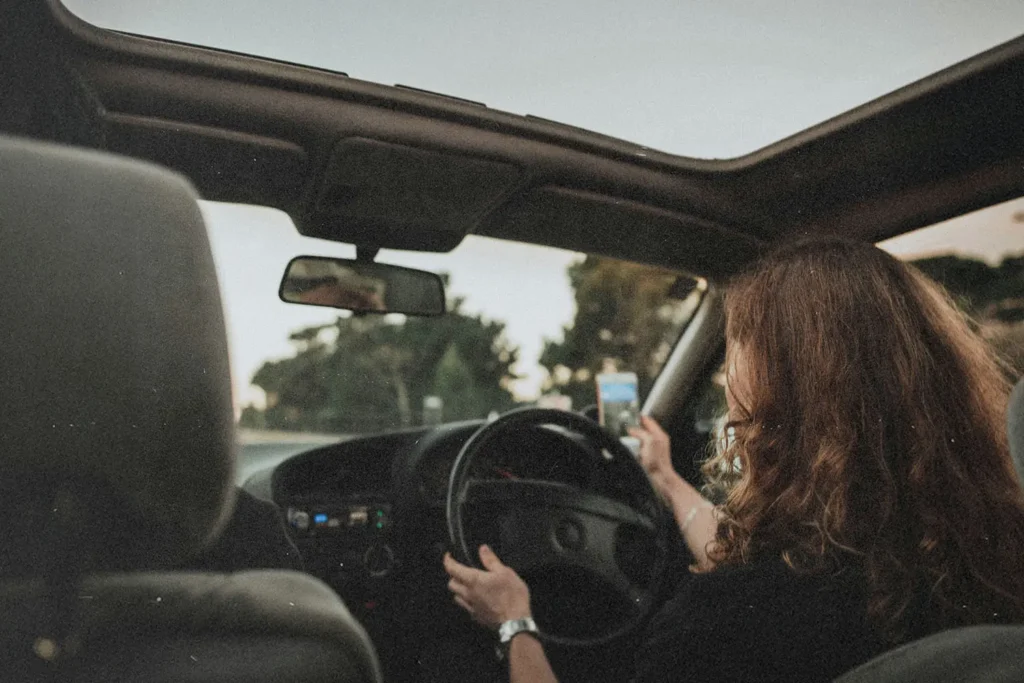 A person drives a car with the roof open, holding a phone in one hand while steering with the other