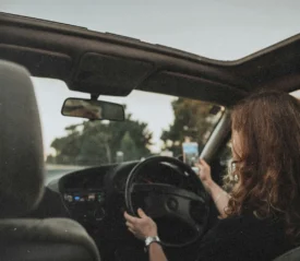 A person drives a car with the roof open, holding a phone in one hand while steering with the other