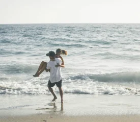 A person carries another through shallow ocean waves along a sunny shoreline