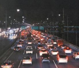 Heavy nighttime traffic on a multi-lane highway, with rows of brake lights stretching into the distance beside a riverside cityscape