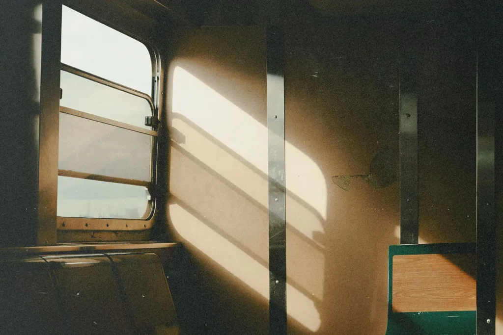 Warm sunlight streams through the window of an old train car, casting long, soft shadows across the beige interior walls and empty seats