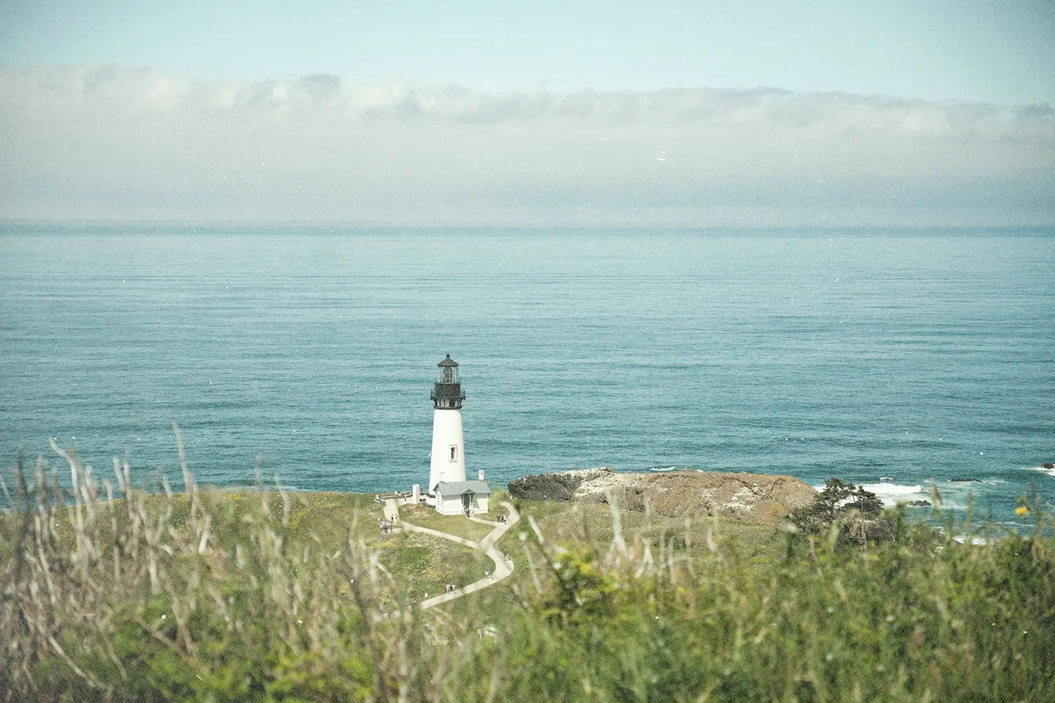 A white lighthouse stands on a grassy cliff overlooking calm blue ocean water, with clouds lining the distant horizon