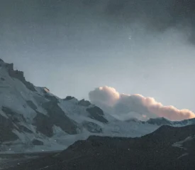 Snow‑covered mountain peaks rise sharply into the sky, illuminated by soft light with a cluster of clouds hovering above