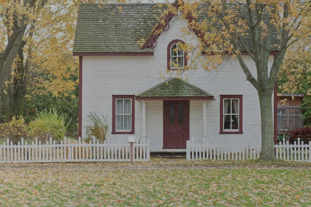 A small white house with red trim sits behind a white picket fence, surrounded by tall trees with golden autumn leaves scattered across the yard