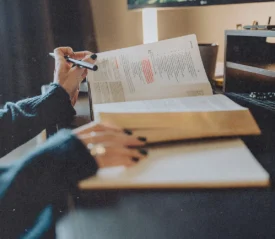 A person studies at a desk with an open book and a Bible, highlighting passages and taking notes in a notebook under soft natural light